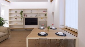 A modern living room with a dining table set for two, a sofa, and a TV on a white wall.