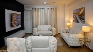 Una sala de estar moderna con un televisor de pantalla plana, dos sofás blancos, una mesa de centro de vidrio y un ventilador de techo.