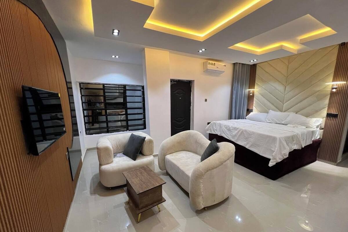 Une chambre moderne avec un grand lit, deux fauteuils, une petite table en bois et un téléviseur fixé au mur.
