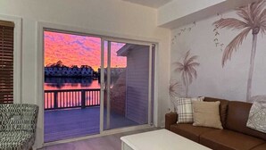 Olohuoneessa on sohva, valkoinen sohvapöytä ja kuviollinen nojatuoli. Siellä on myös lasiliukuovi, joka johtaa parvekkeelle, jolta avautuu näkymä auringonlaskuun.