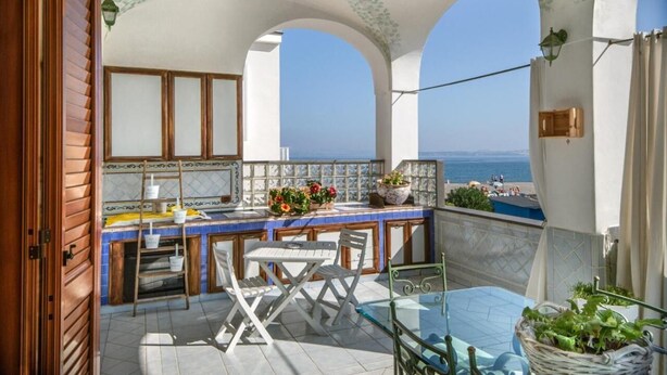 A balcony with a view of the sea, a small table and chairs, a tiled floor, and a window with curtains. - La Finestra Sul Mare (Ischia)