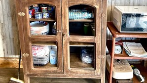 Un armario de madera con estantes abiertos que contienen diversos objetos, una escoba y un televisor al fondo.