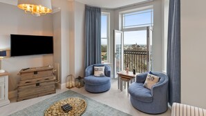 A modern living room with a flat-screen TV, two blue armchairs, a wooden chest of drawers, and a large window offering a city view. - 10 Mansion House - Indoor Pool Sea Views Gym (Tenby)