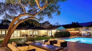 An outdoor pool area with a fire feature, seating, and a view of a house in the background.