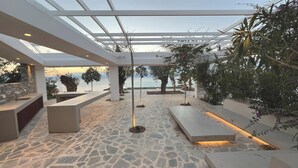Un patio moderno al aire libre con suelo de piedra, pérgola blanca y zonas de estar con vistas a un cuerpo de agua.