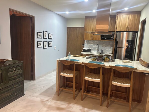 A modern kitchen with wooden cabinets, a stainless steel refrigerator, and a bar area with stools.