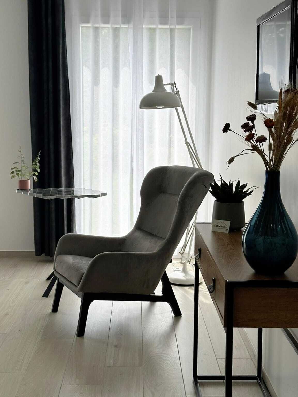 Un salón moderno con un sillón gris, una lámpara de pie, una mesita con un florero de flores secas y una planta en maceta sobre una estantería.