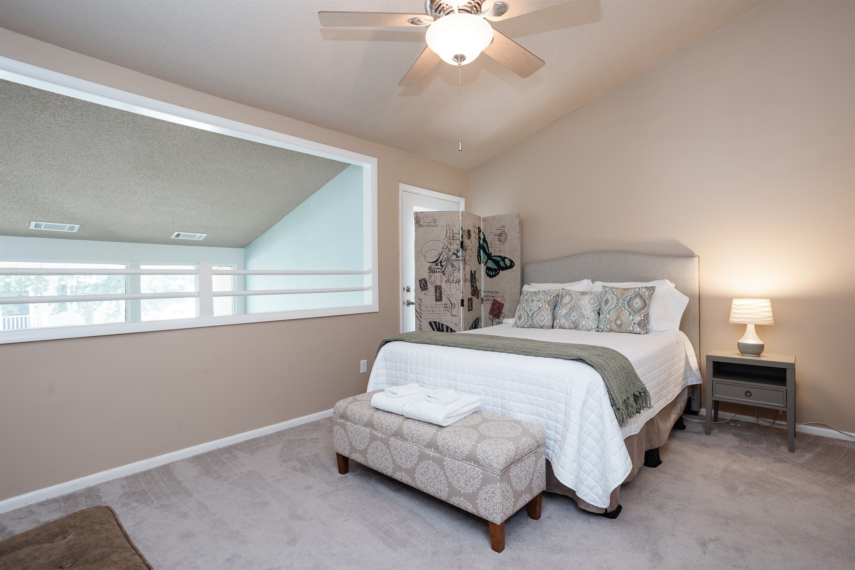 A bedroom with a large window, a ceiling fan, a bed with a headboard, a bedside table with a lamp, and a small bench.