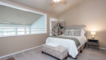 A bedroom with a large window, a ceiling fan, a bed with a headboard, a bedside table with a lamp, and a small bench.