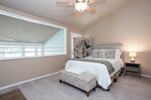A bedroom with a large window, a ceiling fan, a bed with a headboard, a bedside table with a lamp, and a small bench. - Historic District, Walkable w/Free Parking & Patio (Savannah)