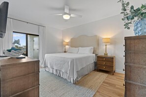 A bedroom with a bed, bedside lamps, a ceiling fan, a dresser, and a view through a door. - Shipwatch Riverside E104 (Pensacola)