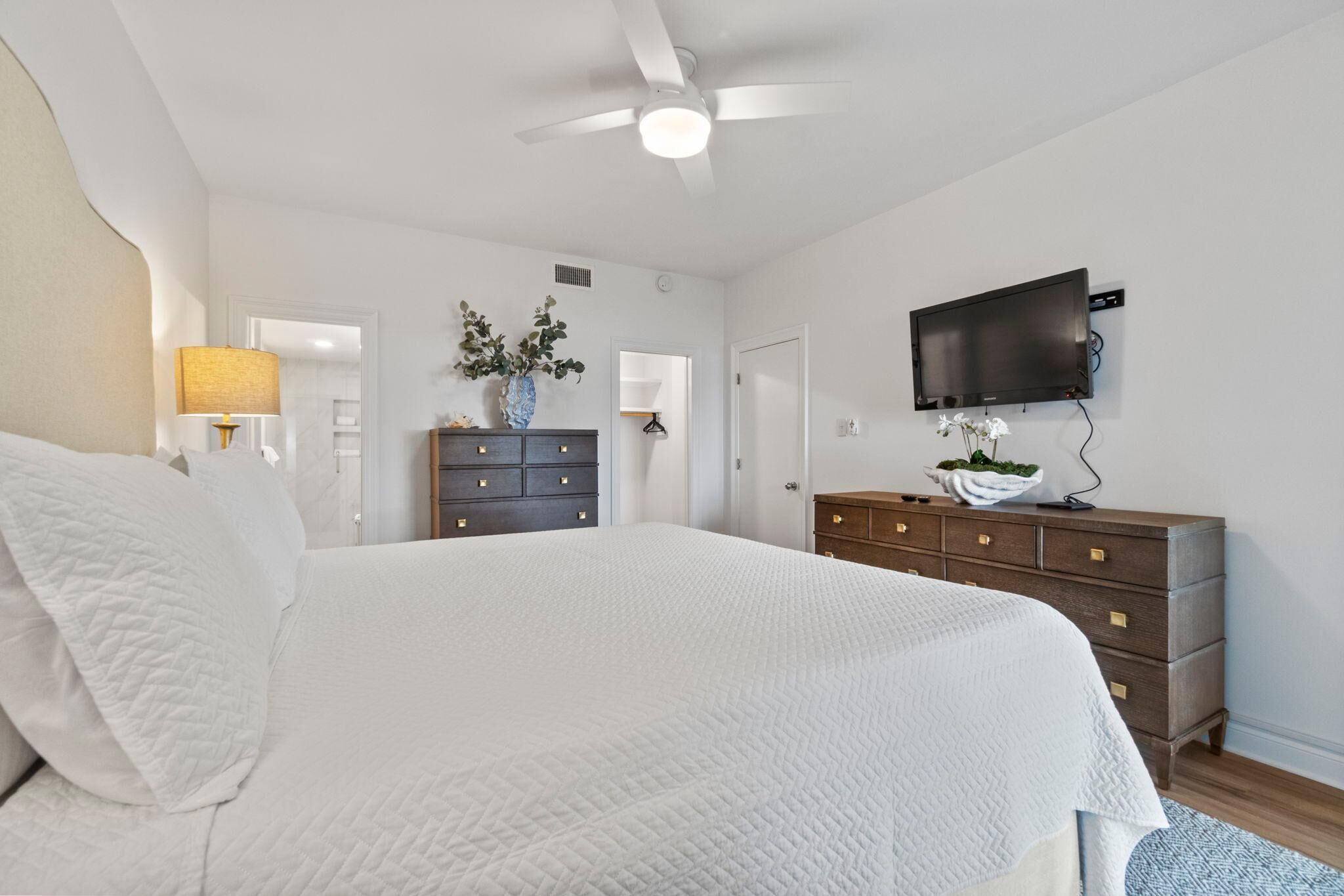 Une chambre avec un grand lit, un ventilateur de plafond, un téléviseur fixé au mur et une commode avec des tiroirs.