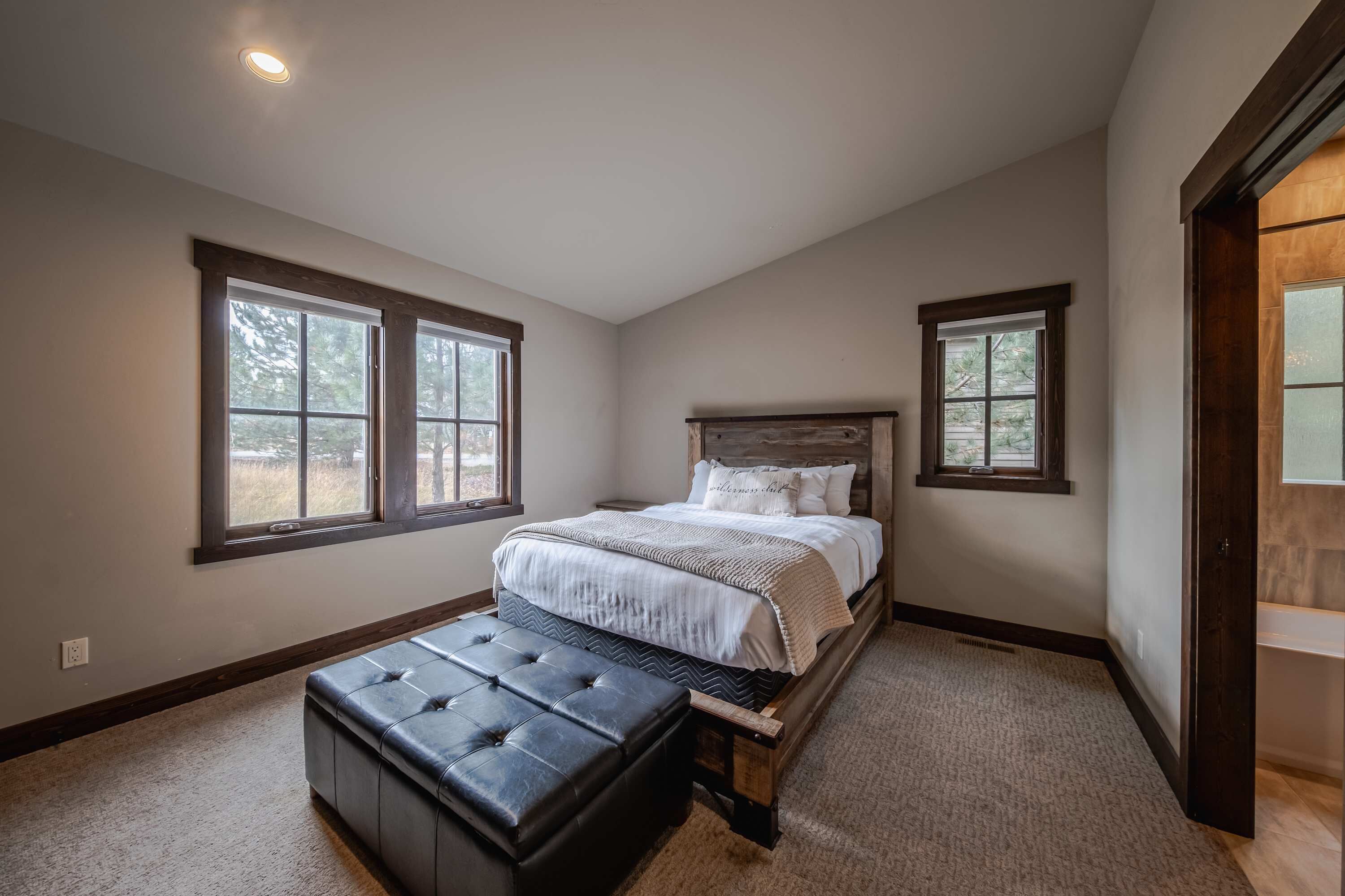 A bedroom with a large bed, a wooden headboard, and a black ottoman. There are two windows providing natural light.