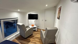 A compact bedroom with a bed, a desk, two chairs, a TV, and a window.