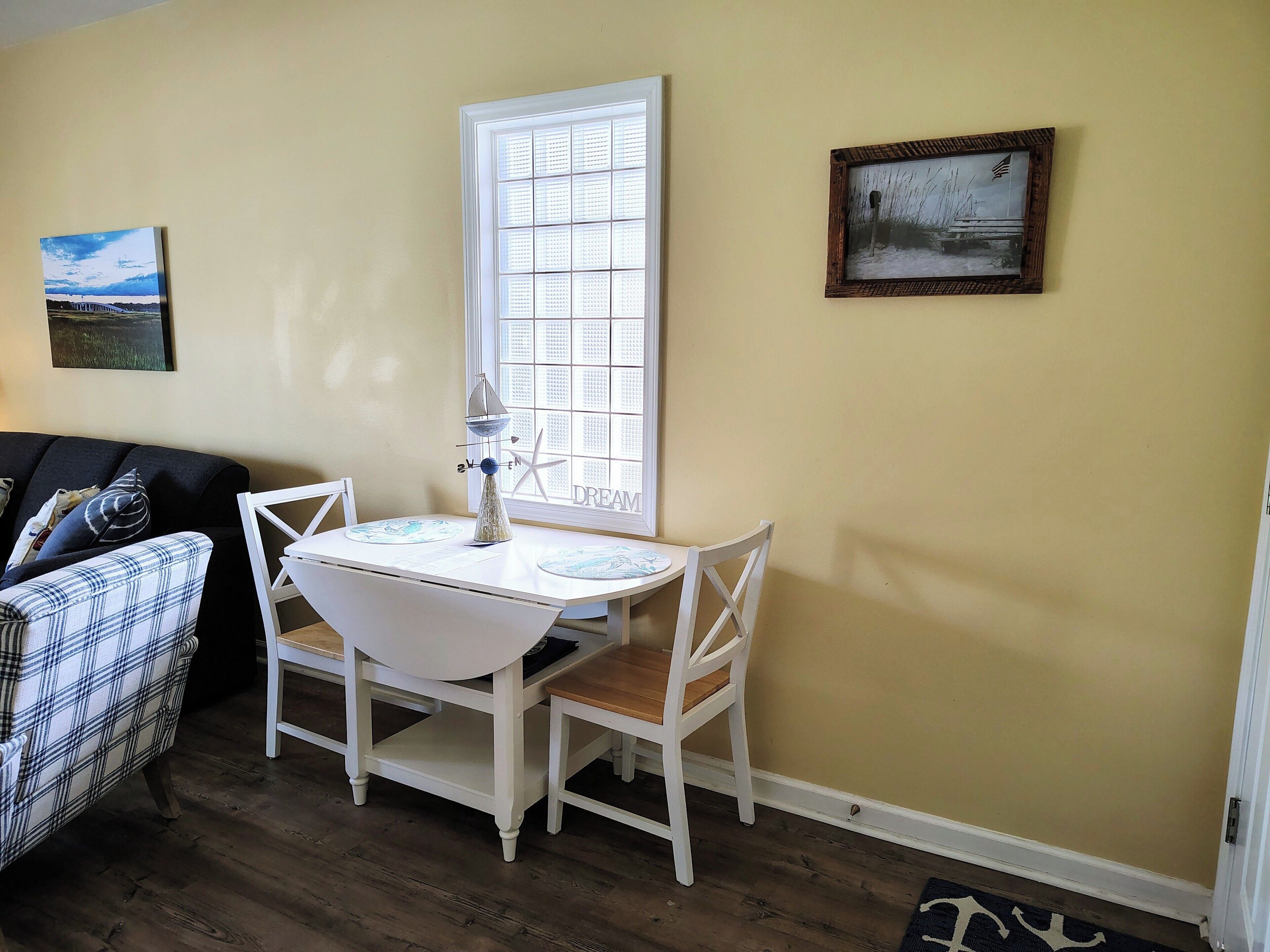 A dining area with a white table and chairs, a window with blinds, and two framed pictures on the wall.
