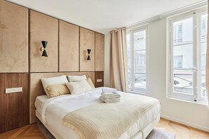 Een slaapkamer met een groot bed, een houten paneel hoofdbord, twee wandlampen, een raam met gordijnen en uitzicht op buiten.