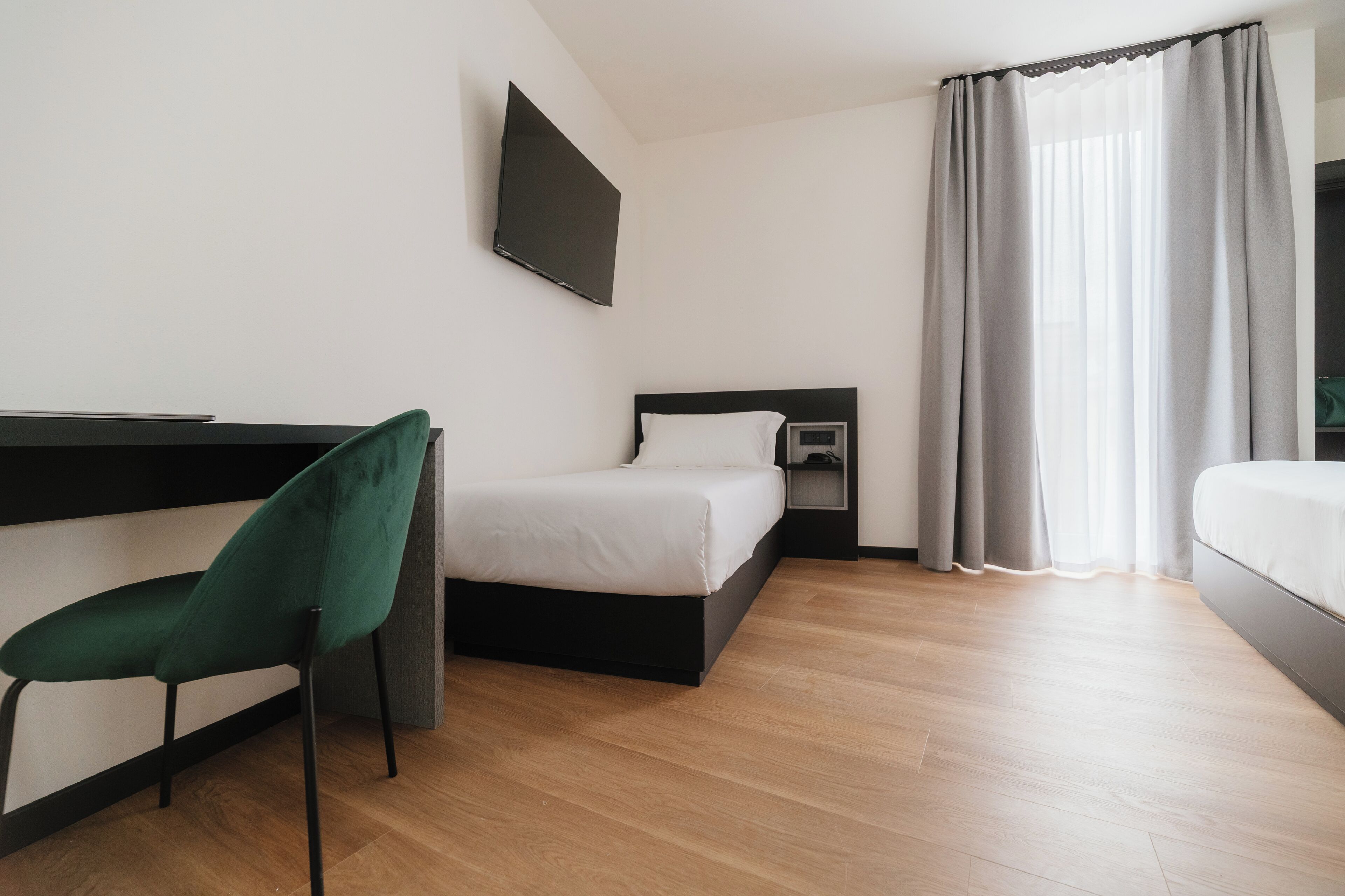A modern hotel room with a bed, a desk, a chair, a TV, and a window with curtains.