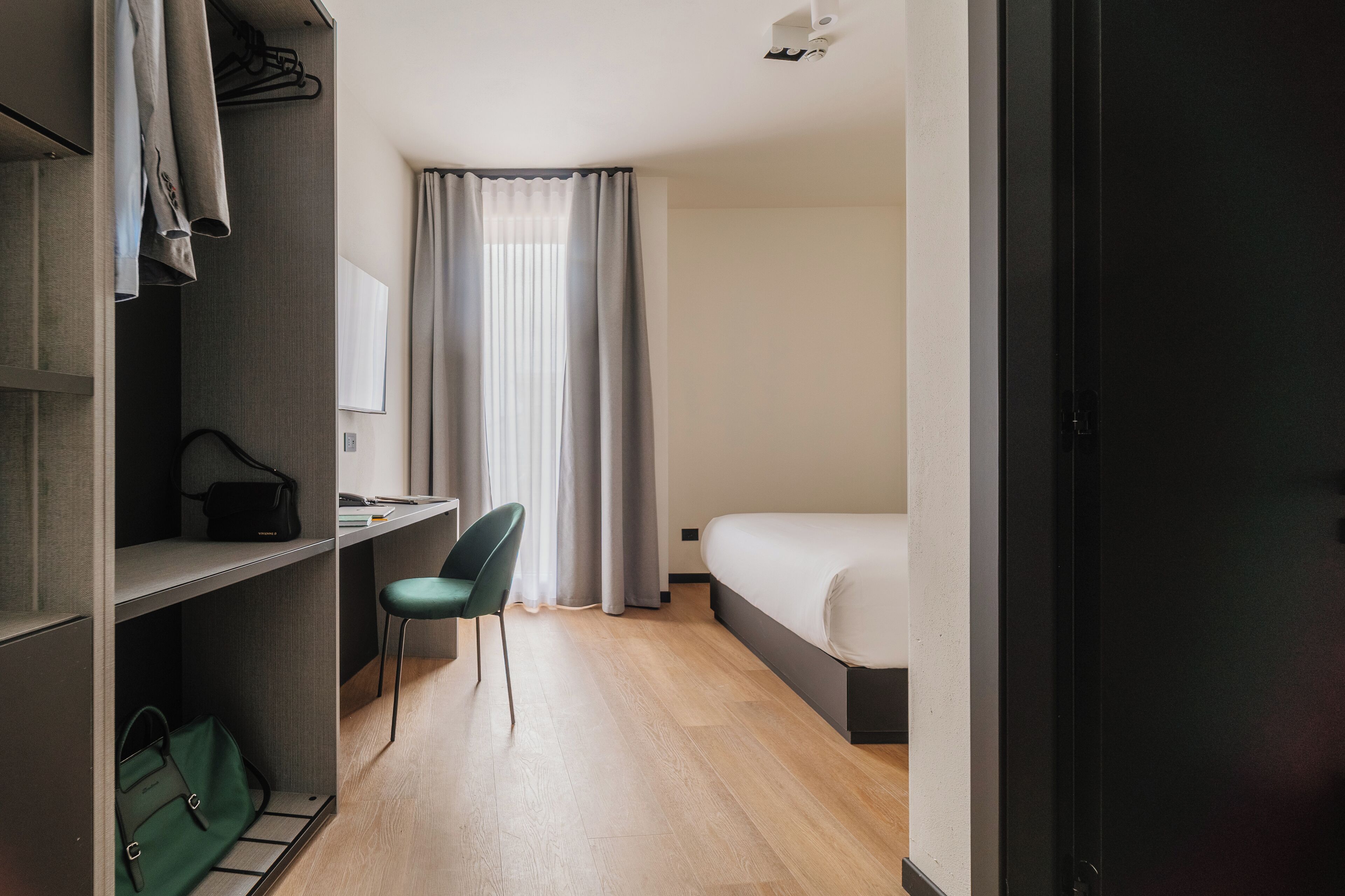 A hotel room with a bed, a desk with a chair, a wardrobe, and a green handbag on the floor.
