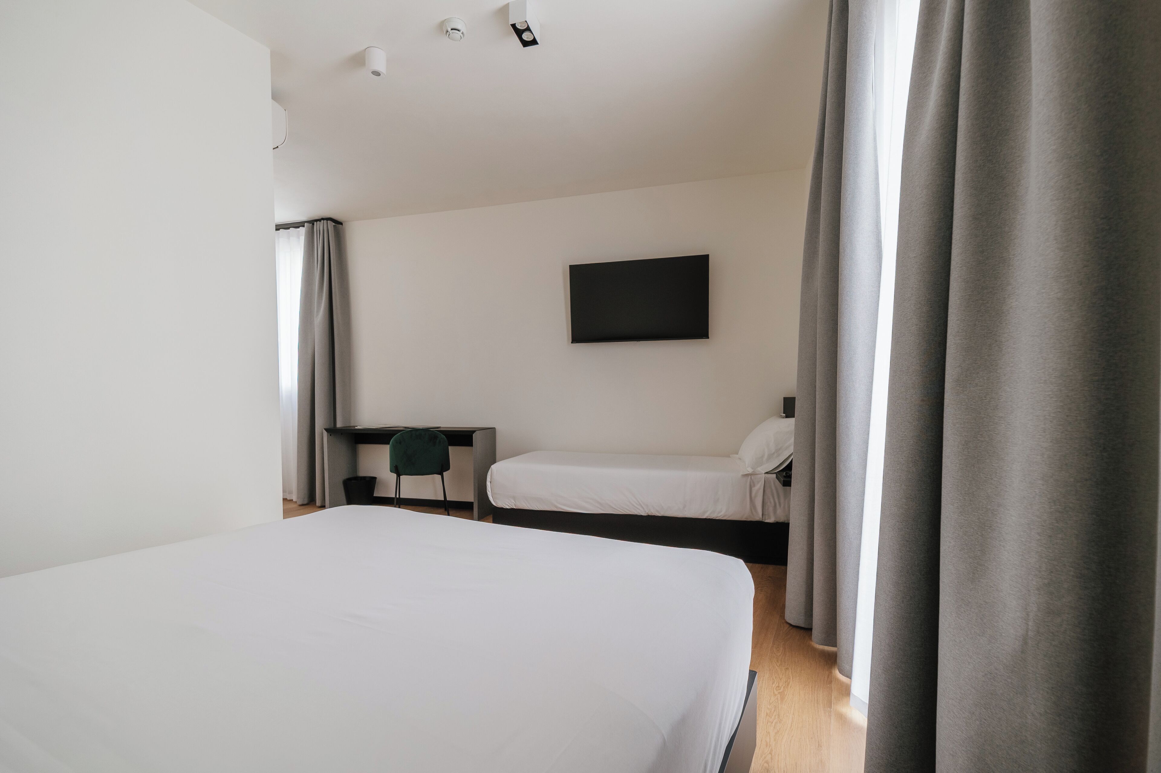 A hotel room with a large bed, a desk with a chair, a TV on the wall, and a window with curtains.