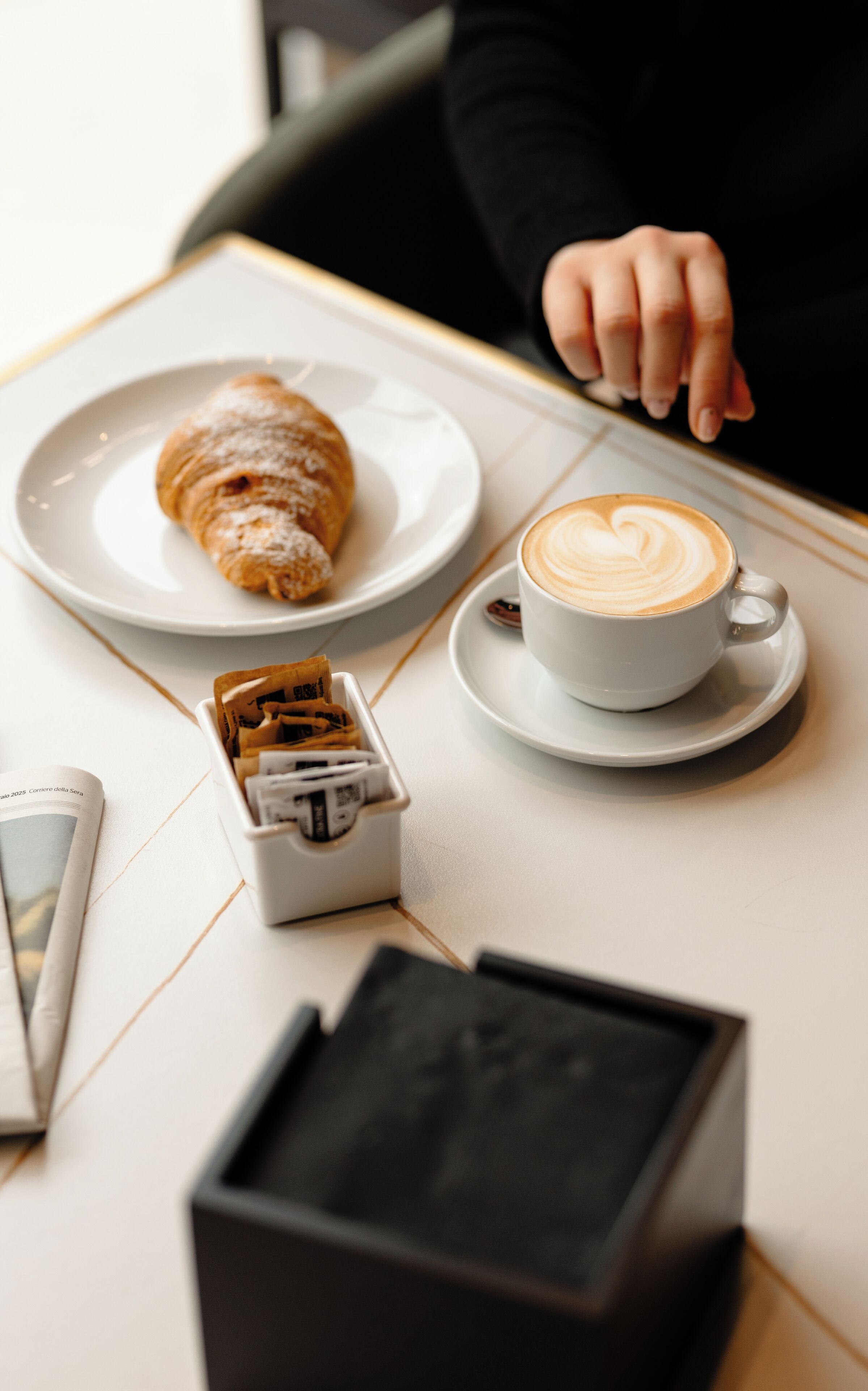 A table with a croissant, a cup of coffee, and a newspaper.