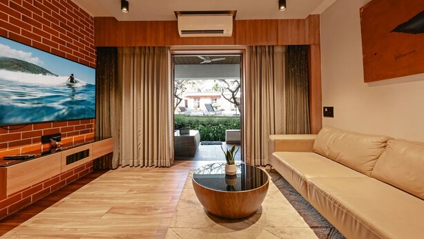 A modern living room with a brick wall, a large flat-screen TV, a wooden coffee table, a beige sofa, and a view of a pool and garden through a glass door. - Charming 1-bedroom apartment in delightful Arpora with WiFi, AC, fitness room (Arpora)