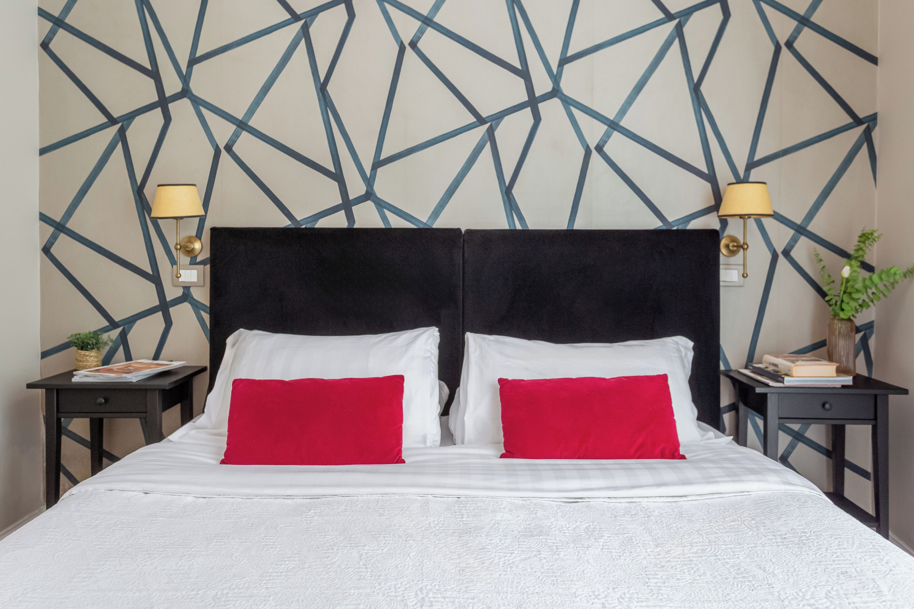 Habitación de hotel con pared de diseño geométrico, cabecera tapizada en negro, ropa de cama blanca y dos almohadas rojas. Hay dos mesitas de noche, una con lámpara y otra con una planta pequeña.