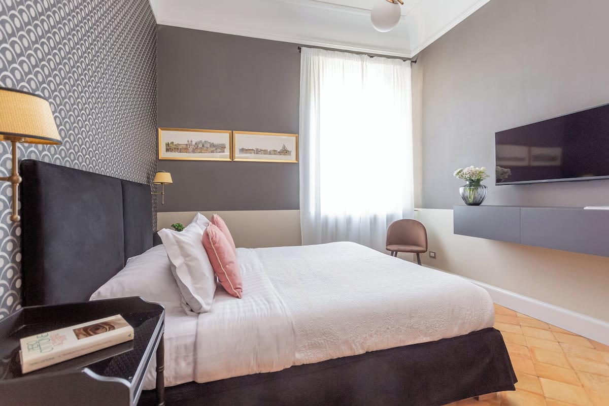 A modern bedroom with a large bed, a flat-screen TV, a desk with a lamp, and framed pictures on the wall.