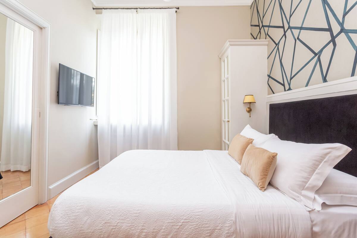 A neatly made bed with white linens and beige pillows, a flat-screen TV mounted on the wall, and a geometric-patterned wall above the bed.