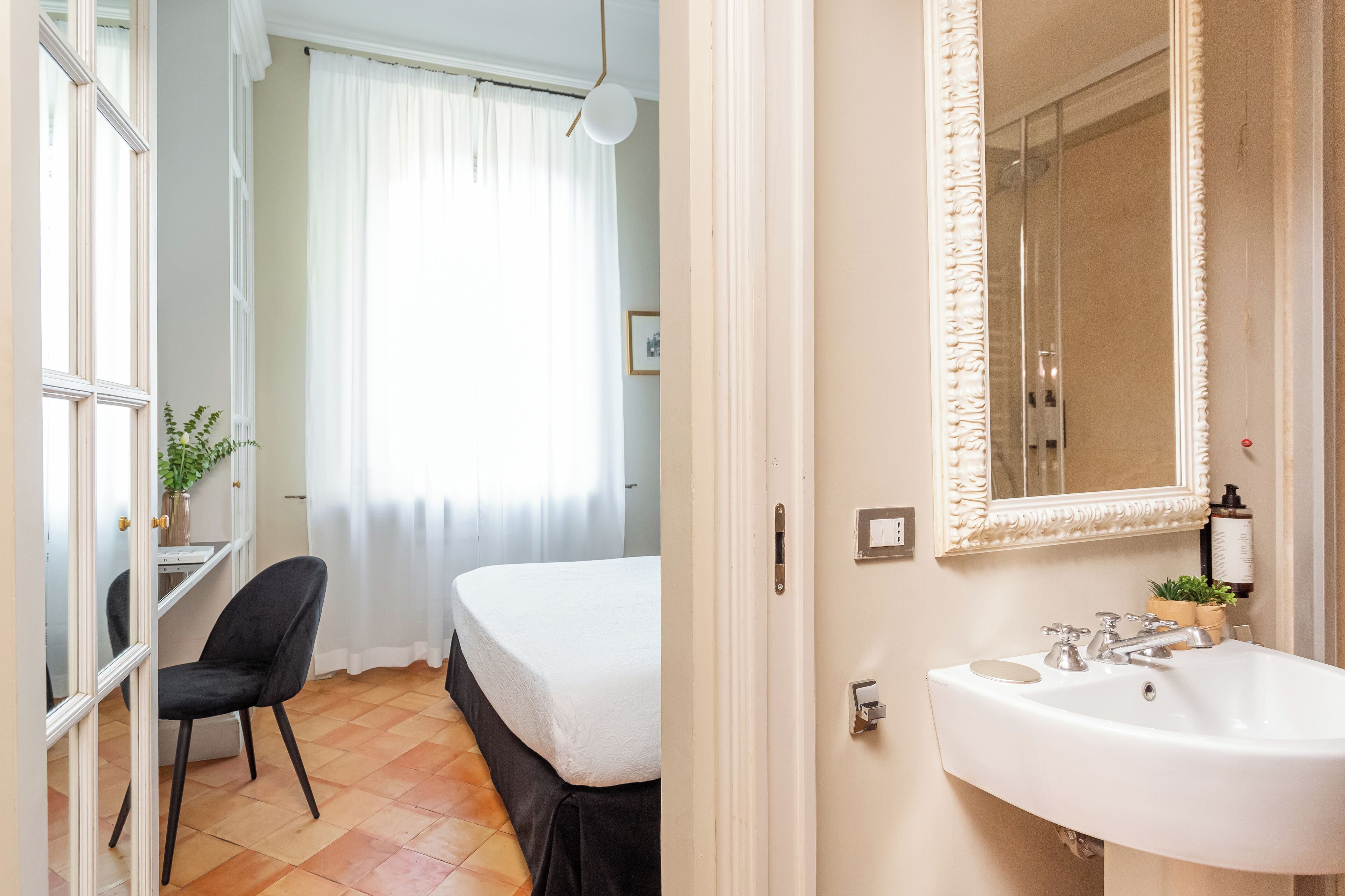 Habitación de hotel con cama, una silla, escritorio, un espejo grande y baño con lavamanos y espejo.