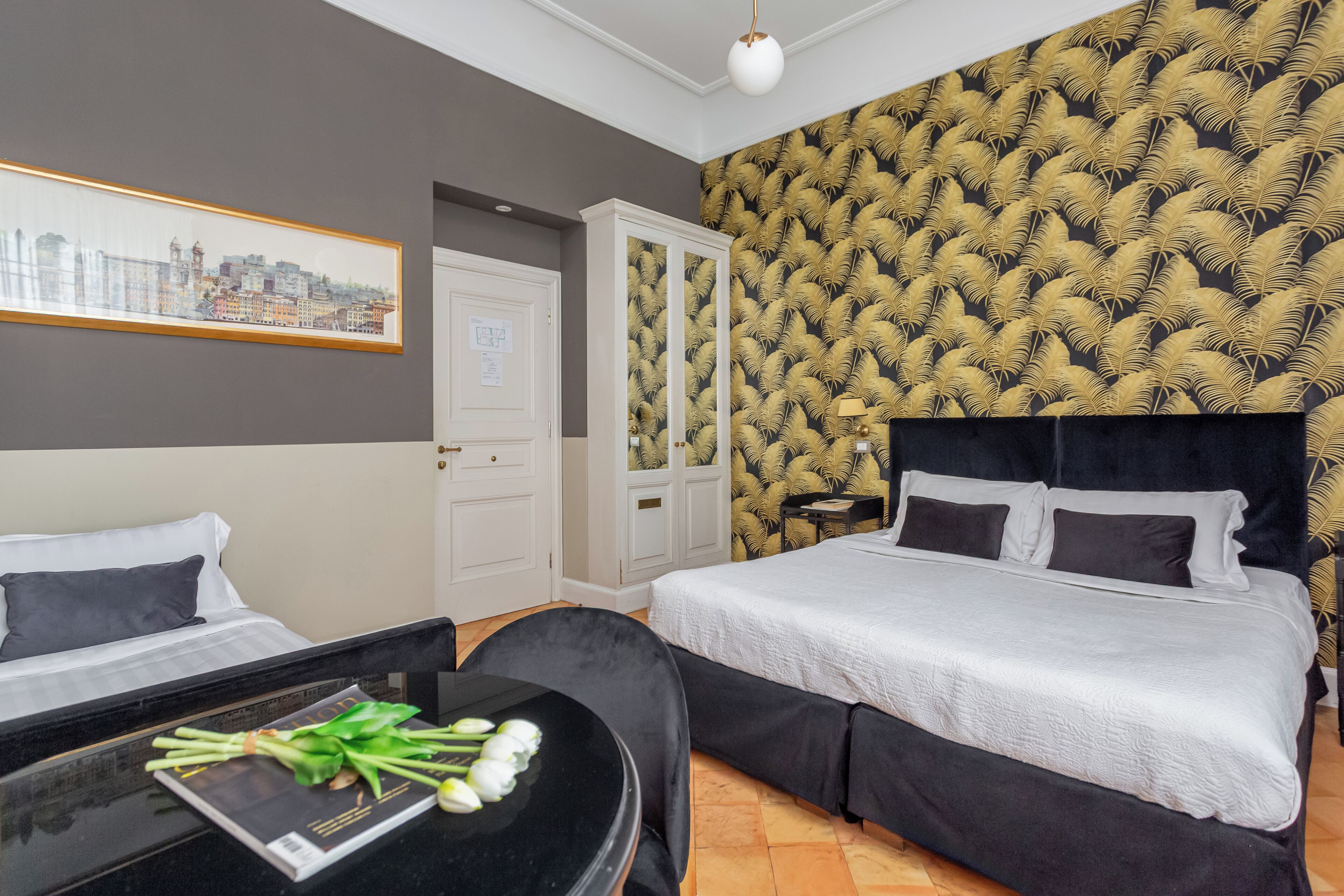 Une chambre d’hôtel avec un lit, un bureau et une chaise. Un tableau représentant un paysage urbain est accroché au mur, et le papier peint est à motifs.