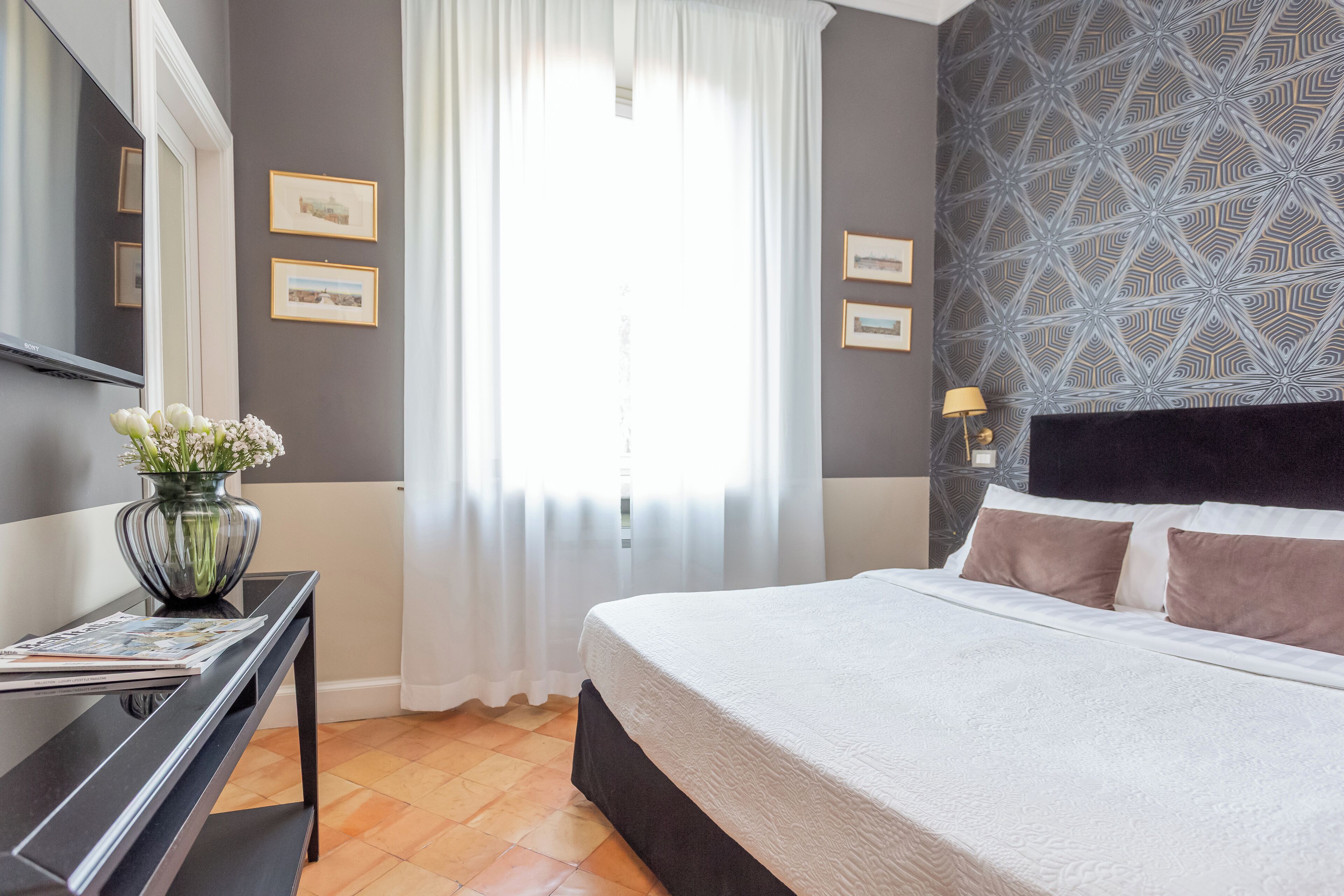 A bedroom with a bed, a desk, a vase with flowers, and a window with curtains.