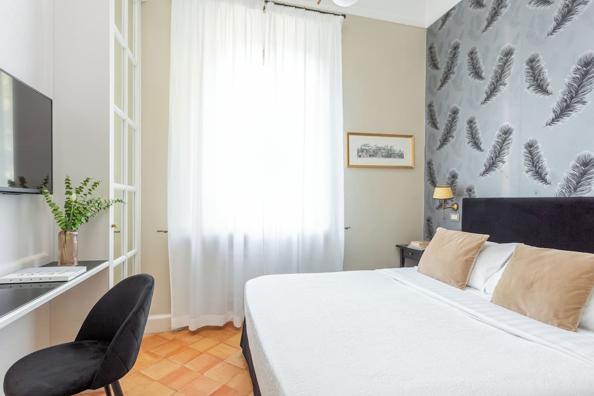 A bedroom with a bed, a desk, a chair, a TV, and a window with curtains.