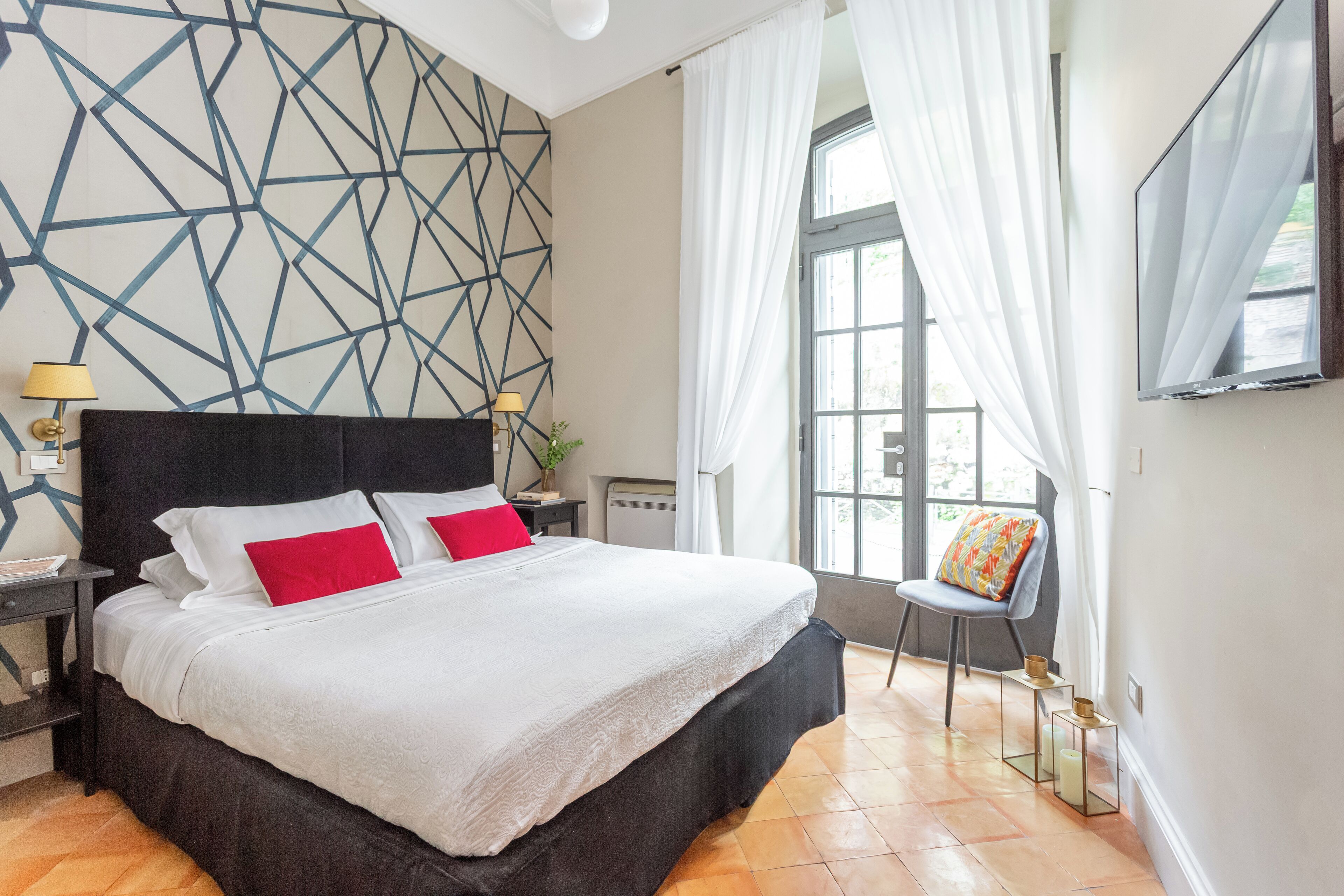 Una habitación de hotel moderna con una cama grande, un muro con patrón geométrico, una ventana con cortinas translúcidas y un televisor de pantalla plana montado en la pared.