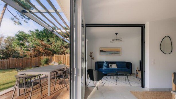 Un salon moderne avec un canapé bleu, une table à manger et des chaises, et une vue sur l’extérieur grâce à de grandes portes vitrées.