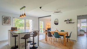 Une cuisine moderne dotée d’un îlot central, de tabourets de bar, d’une table à manger avec des chaises, de luminaires suspendus et d’une vue à travers la porte vitrée.