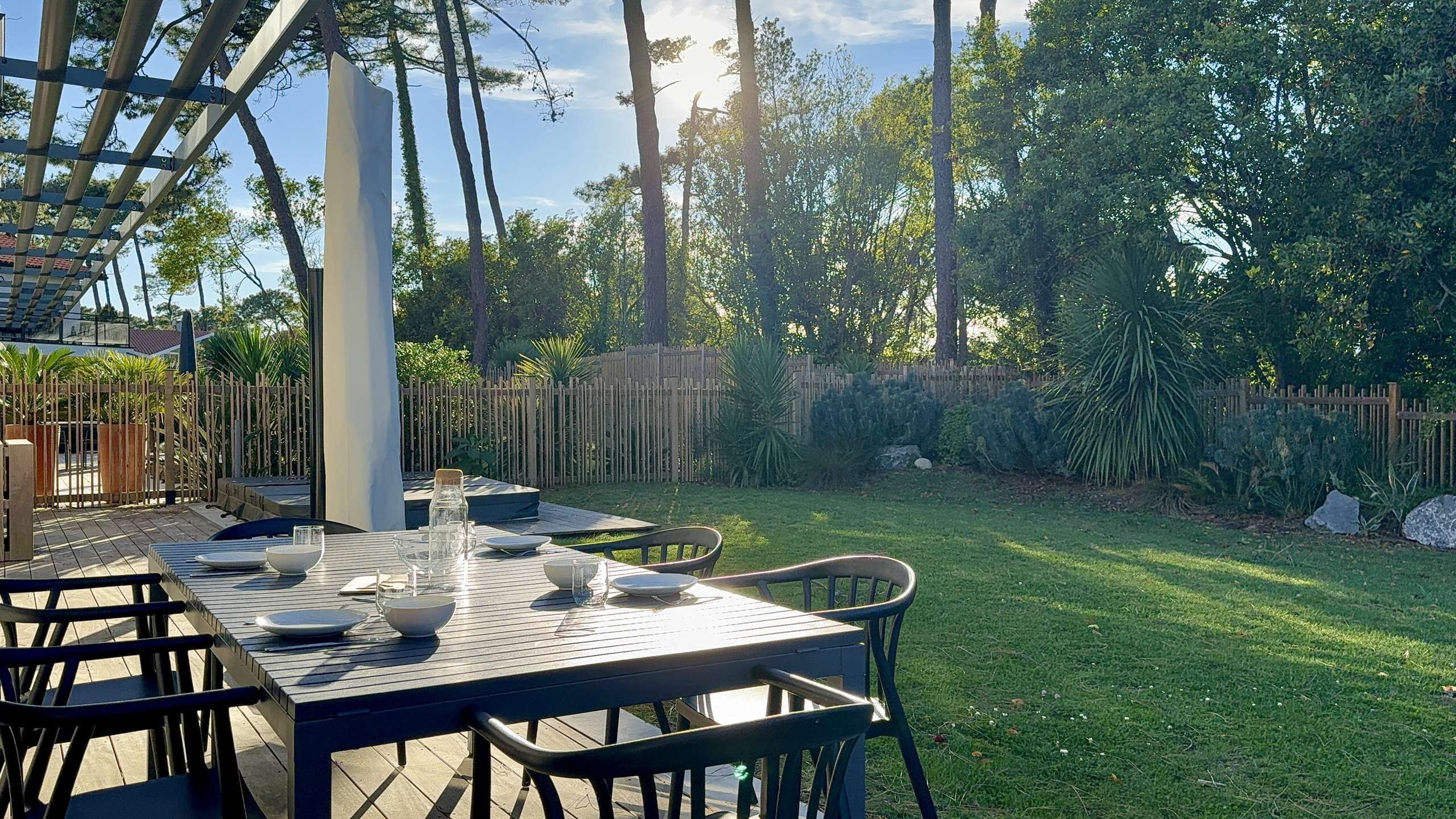 An outdoor dining area with a table set for four, surrounded by chairs, in a garden with a wooden fence and trees.