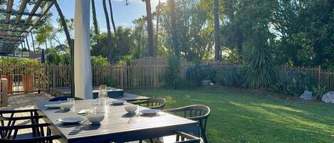 An outdoor dining area with a table set for four, surrounded by chairs, in a garden with a wooden fence and trees.