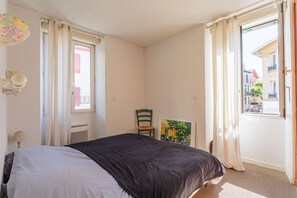 Une chambre à coucher avec un lit, une chaise, un tableau et une vue sur la rue, visible par une porte ouverte.