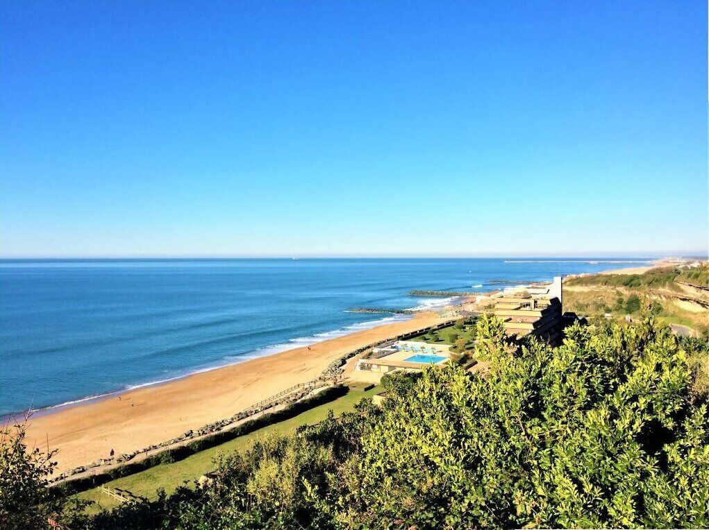 A coastal view with a beach, a swimming pool, and a white building in the distance.