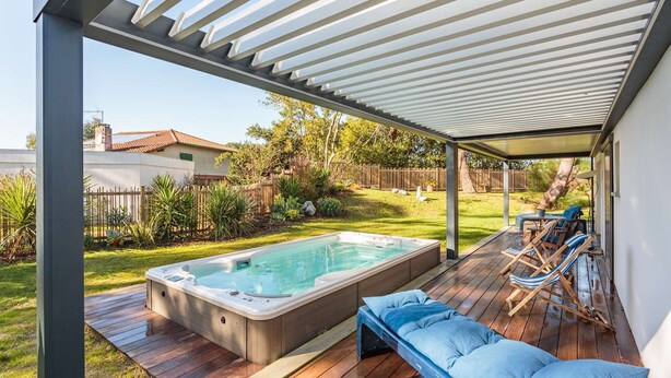 A hot tub under a covered patio with a wooden deck, surrounded by a grassy yard and a wooden fence.
