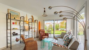 A modern living room with a sofa, armchairs, a glass coffee table, a bookshelf, and a large window.