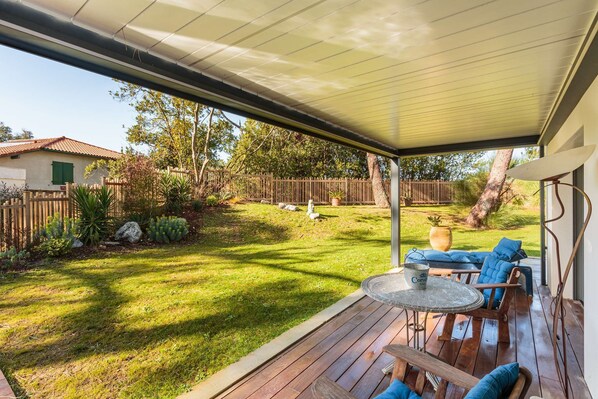 A covered patio with a table and chairs, a view of a backyard with a wooden fence, and a house in the background.