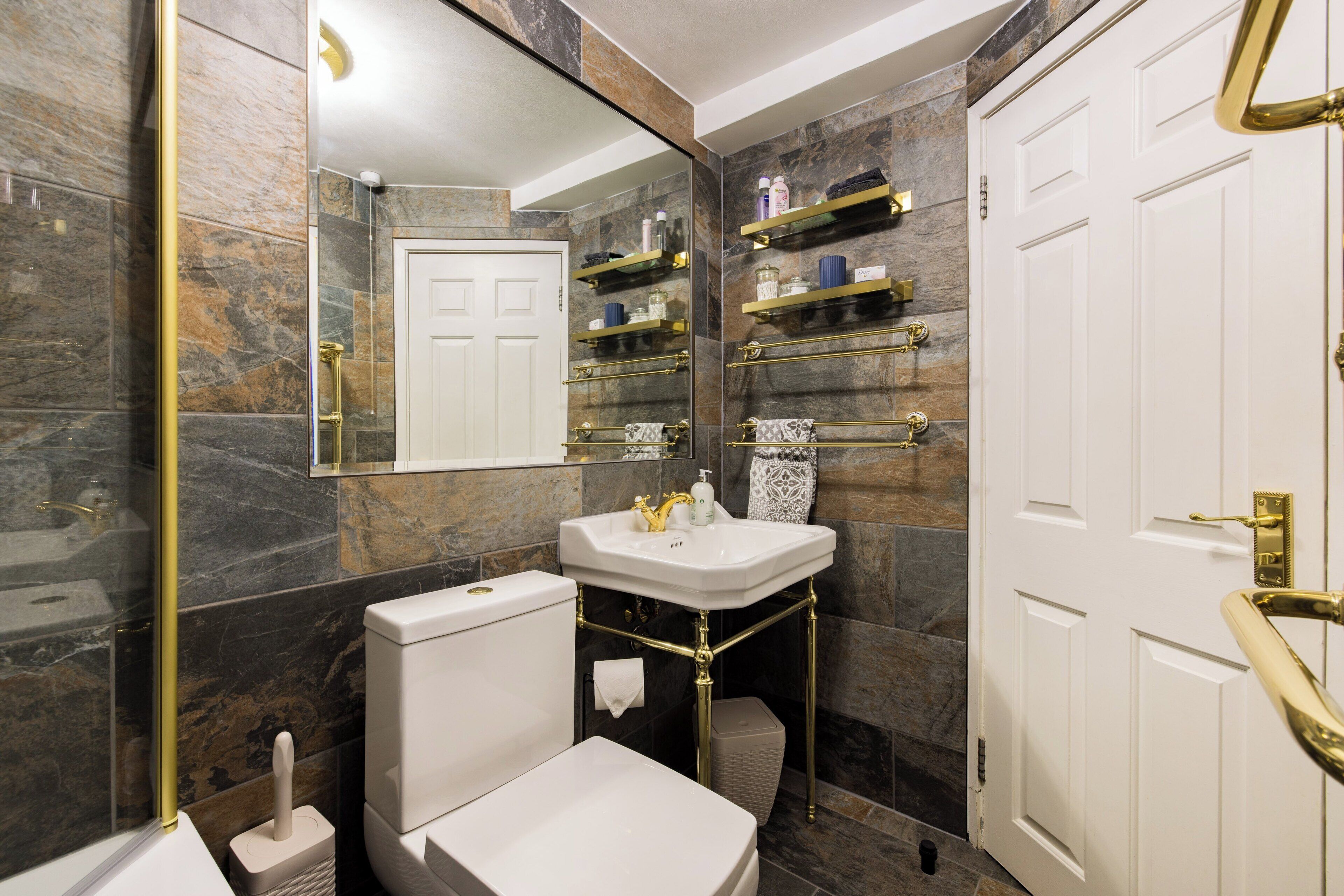 Un baño moderno con inodoro blanco, lavamanos y espejo. Hay estantes sobre el lavamanos y una puerta con pomo dorado.