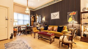 A well-lit living room with a yellow sofa, wooden chairs, a coffee table, and a chandelier. - Large 4-bed 8-guests W/patio 1841 House Paddington (London)