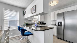 A modern kitchen with white cabinets, a stainless steel refrigerator, a bar area with blue stools, and a window with blinds.