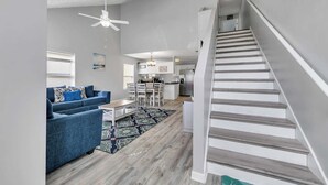 A modern living room with a blue sofa, a dining area, a kitchen, and a staircase.