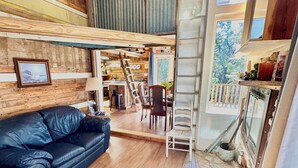 Un salon chaleureux avec un canapé en cuir noir, un plancher en bois, une table à manger avec des chaises, un escalier menant à une mezzanine et une vue sur l’extérieur.