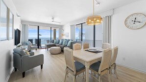 A modern living room with a dining area, large windows, and a view of the ocean. - 🌴🌊Romar Place 806: Beautiful Corner Unit | Beachfront (Orange Beach)