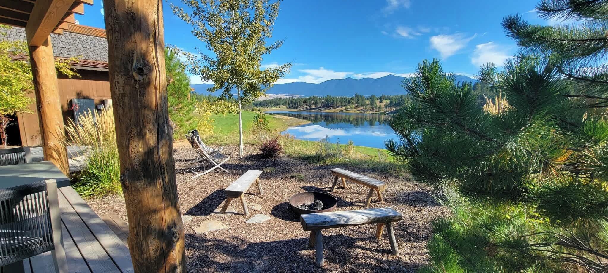 An outdoor seating area with wooden benches and a fire pit, overlooking a lake and mountains.
