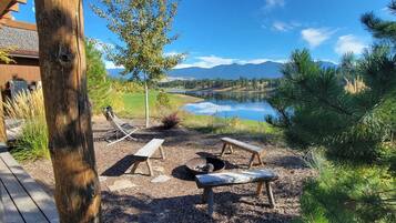 An outdoor seating area with wooden benches and a fire pit, overlooking a lake and mountains.